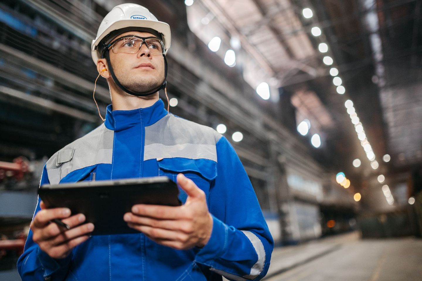 A maintenance engineer in safety gear reading a tablet on the floor of a large industrial hall
